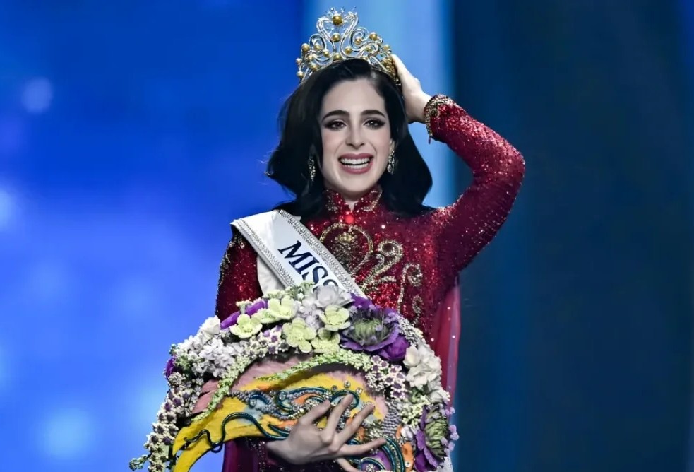 A woman wearing a jeweled crown and a red embellished gown smiles while holding a large floral bouquet and wearing a “Miss” sash on stage during a pageant ceremony.