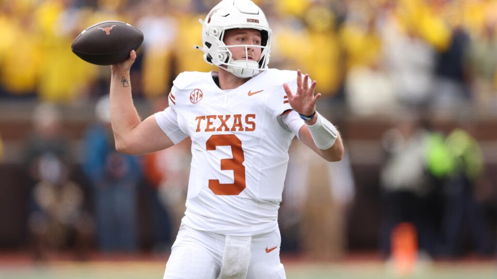 A Texas Longhorns quarterback wearing a white uniform prepares to throw a pass during a game, holding the football mid-motion against a blurred stadium crowd.