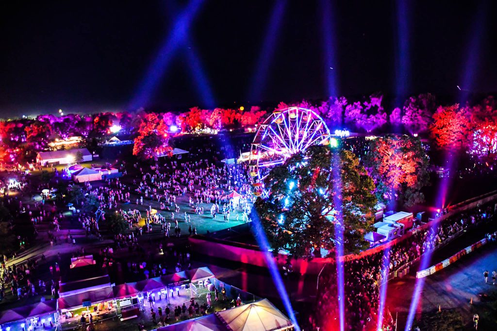 Nighttime aerial view of Bonnaroo 2026 festival grounds with a glowing Ferris wheel, colorful lights, and large crowds gathered across the field.