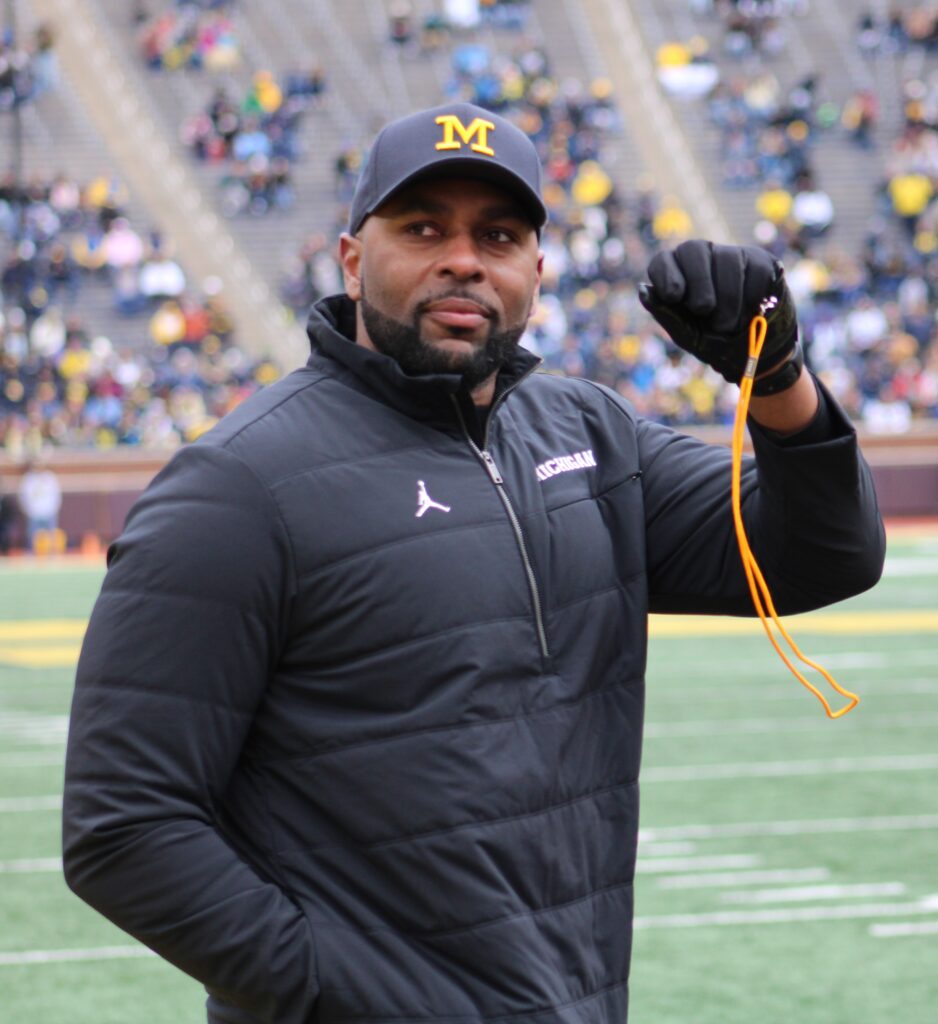 Sherrone Moore: Man wearing a Michigan cap and black athletic jacket standing on a football field holding a whistle